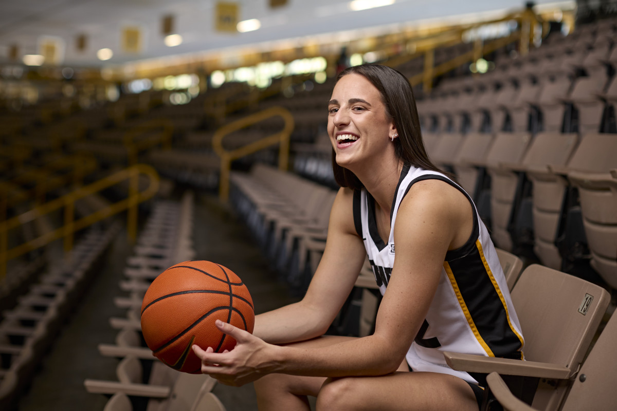 Caitlin Clark smiles and holds a basketball while posing for a portrait in the stands.