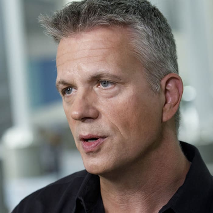 A middle-aged white man with short silver hair is sitting in an office with a blurred-out background. He wears a solemn expression on his face