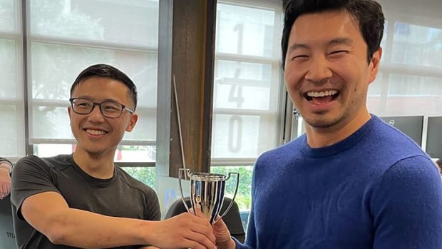 Simu Liu being handed a trophy for winning in a video game.