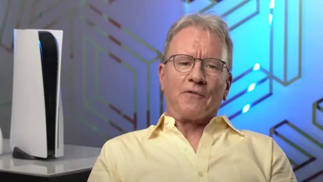 A white man with short blonde hair, wearing thin-rimmed black glasses and a yellow collared shirt, is sitting in front of a table with a PS5 console on it.