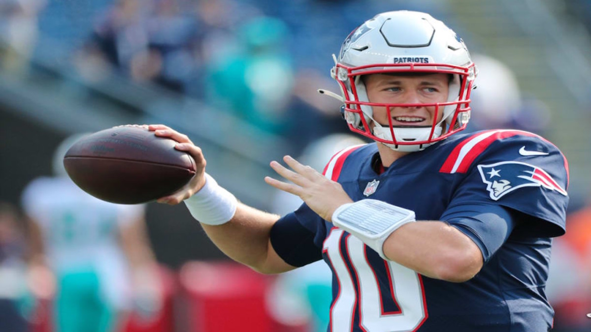 Mac Jones, a white man wearing a silver NFL helmet and navy-blue jersey, is preparing to throw a football in his right hand. He looks ahead with a smile on his face.