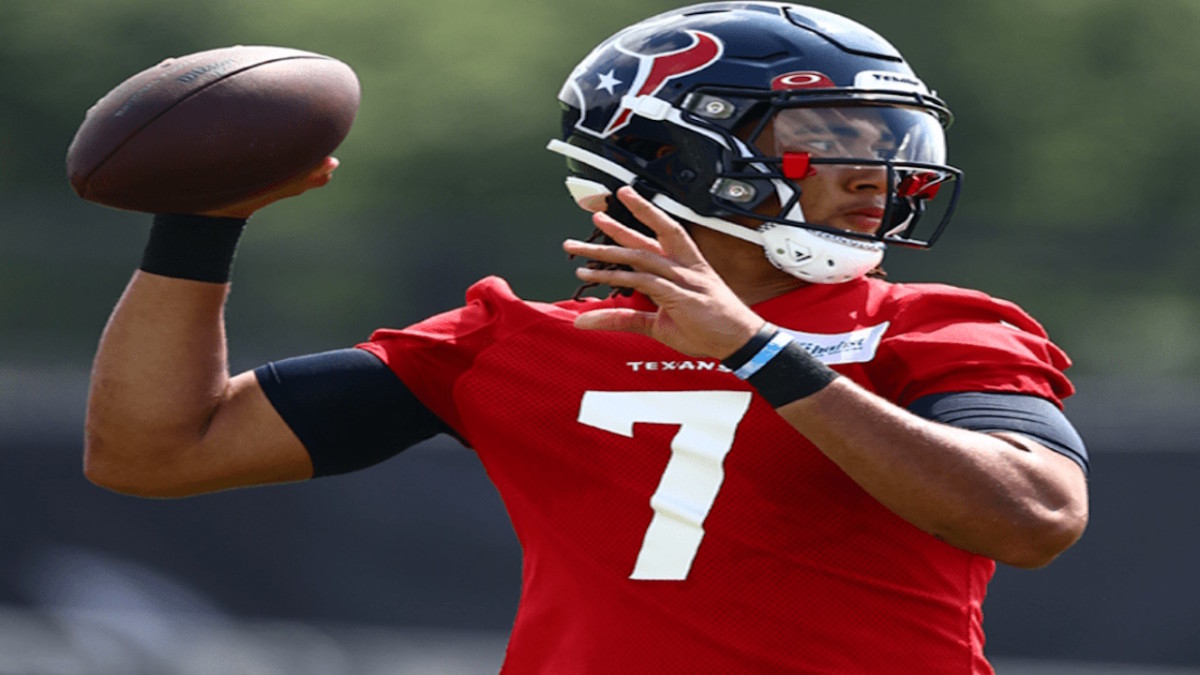 CJ Stroud, a Black man wearing a blue NFL helmet and wearing a red jersey, is throwing a football with his right hand.